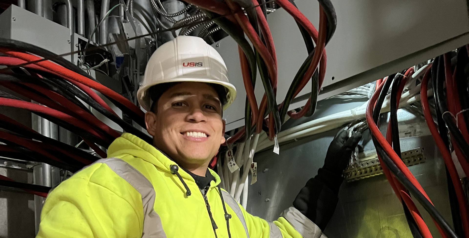 USIS engineer in a hard hat and safety vest stands next to a large electrical panel, ensuring safety.