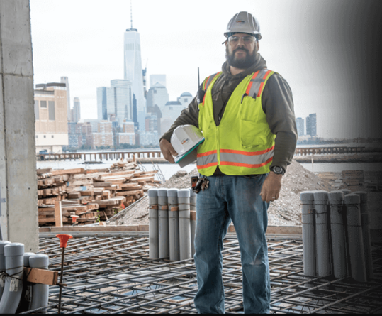 USIS supervisor/engineer with engineering gear standing on a construction site with helmet in hand.