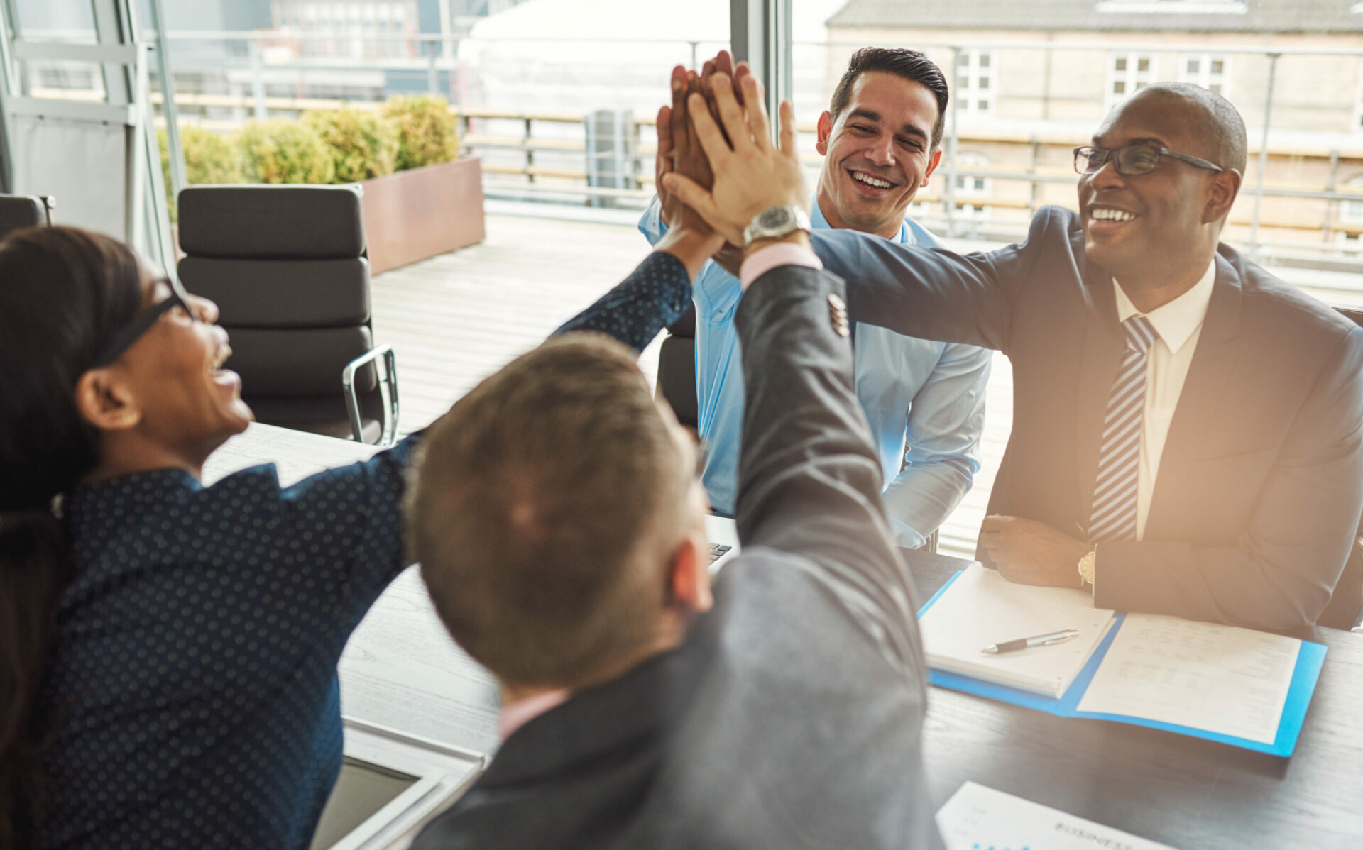 A group of individuals sitting in a meeting room bringing hands together for a high five.